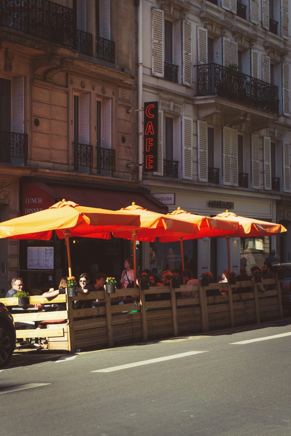 People converse at tables under large umbrellas on a restaurant patio.